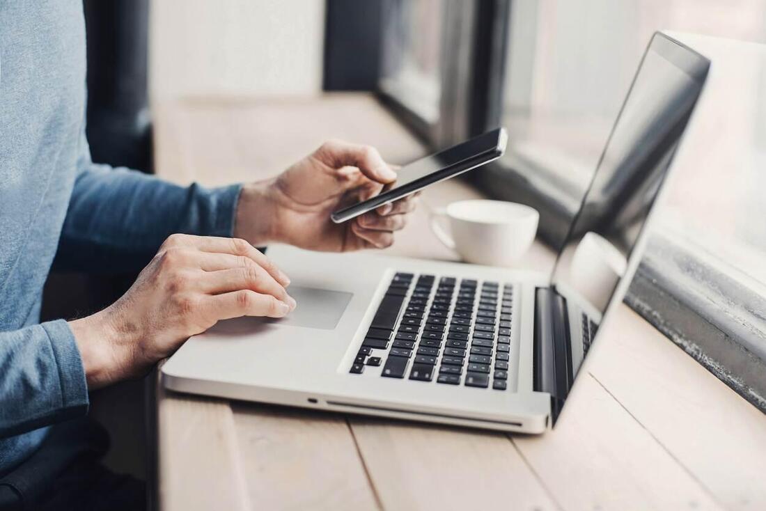 Man zit achter laptop met een mobiel in de linkerhand.